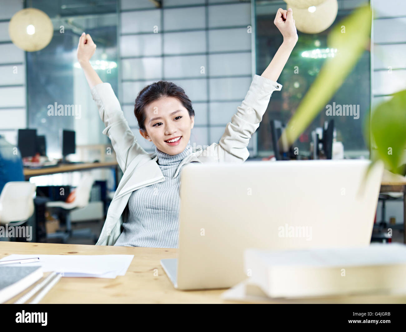 Young Asian business woman celebrating with arms raised in office. Banque D'Images