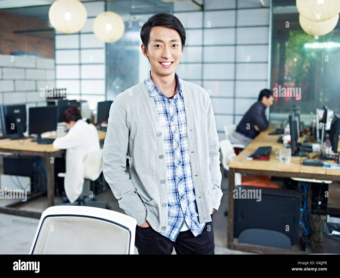 Certains jeunes entrepreneur asiatique debout dans propre entreprise looking at camera les mains dans les poches en souriant. Banque D'Images
