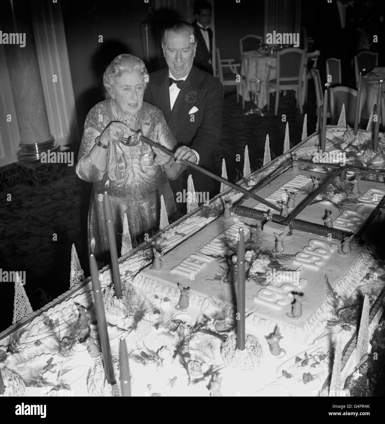 Agatha Christie, auteur de la pièce record 'The Mousetrap', utilise une épée pour couper un gâteau d'anniversaire, pesant une demi-tonne, lors d'une fête à l'Hôtel Savoy pour célébrer le 10e anniversaire de la course de sa pièce 'The Mousetrap' au Théâtre de l'Ambassadeur.Peter Saunders l'aide, qui présente la pièce.Le gâteau avait un piège géant de souris de massepain sur lui et 10 bougies.La fête, organisée par Agatha Christie et M. Saunders, a réuni 1,000 invités, dont la plupart des stars du monde du divertissement et de nombreux acteurs et actrices qui ont figuré dans la pièce. Banque D'Images
