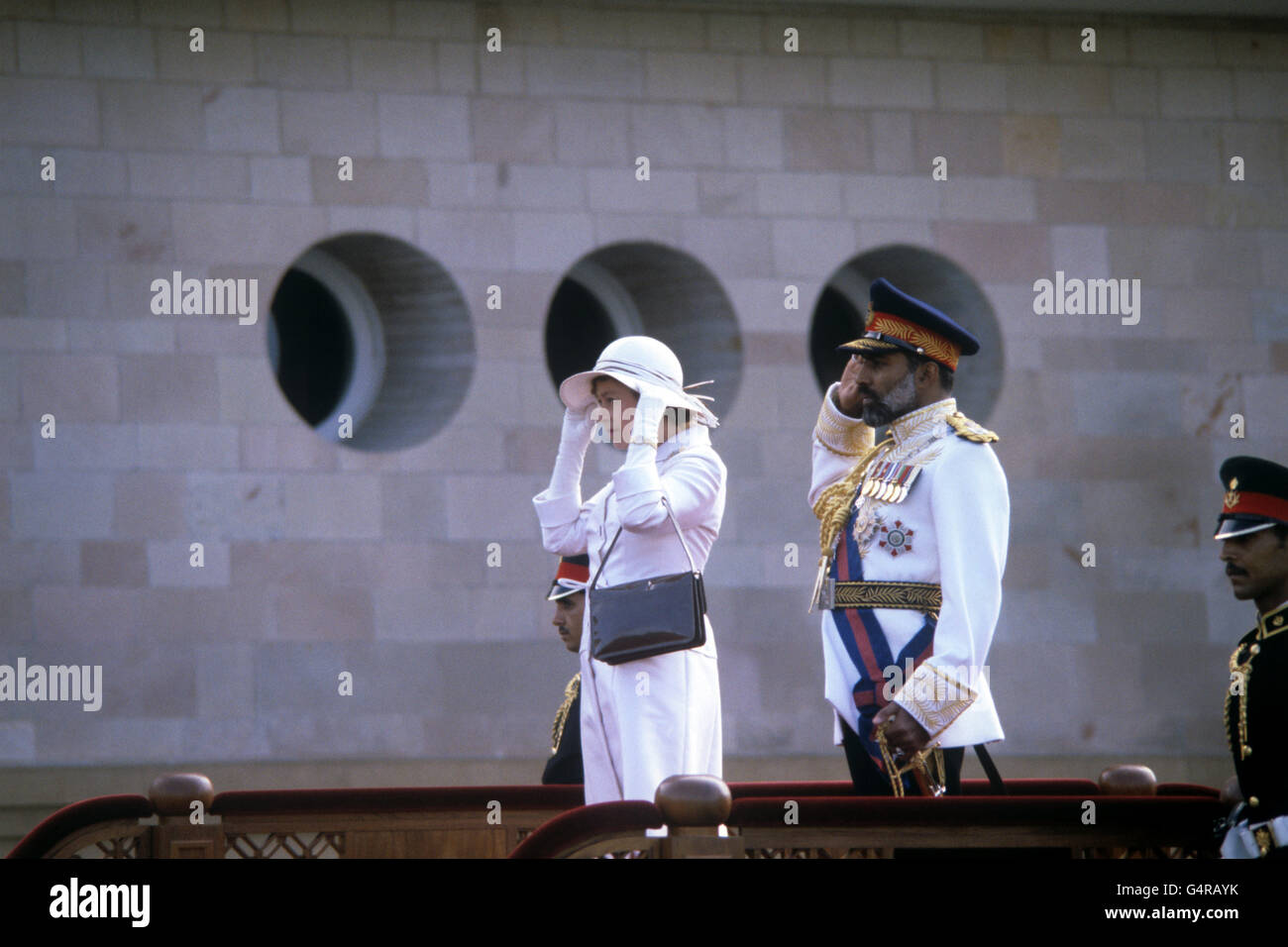 La reine Elizabeth II tient sur son chapeau comme elle se tient aux côtés du Sultan d'Oman, Sultan Qaboos bin a dit, comme ils prennent le salut pendant le jeu de l'hymne national dans le domaine du palais de Muscat. Banque D'Images