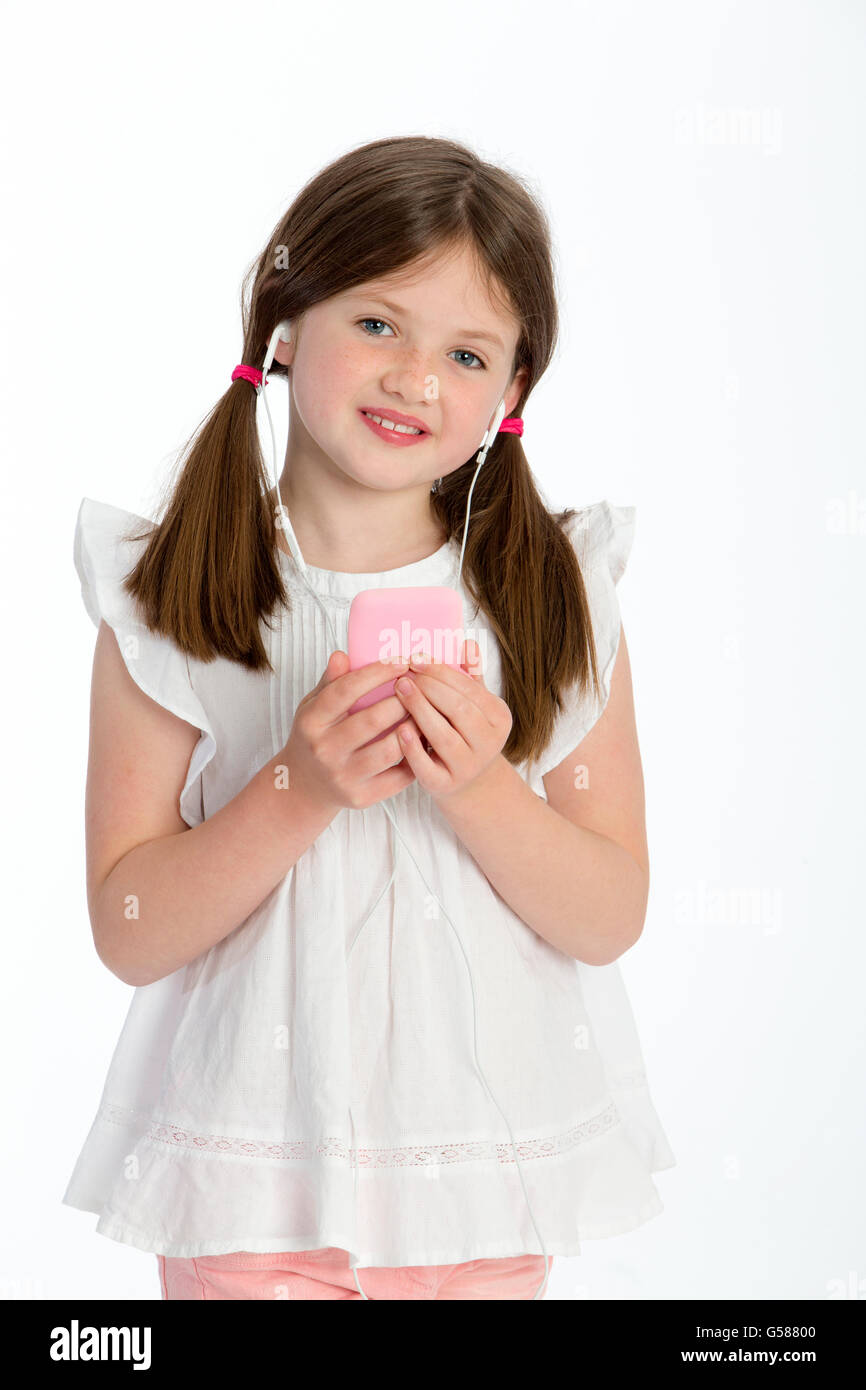 Petite fille l'écoute de musique au casque sur un smartphone. Elle sourit à la caméra et se tenir contre un bac blanc Banque D'Images