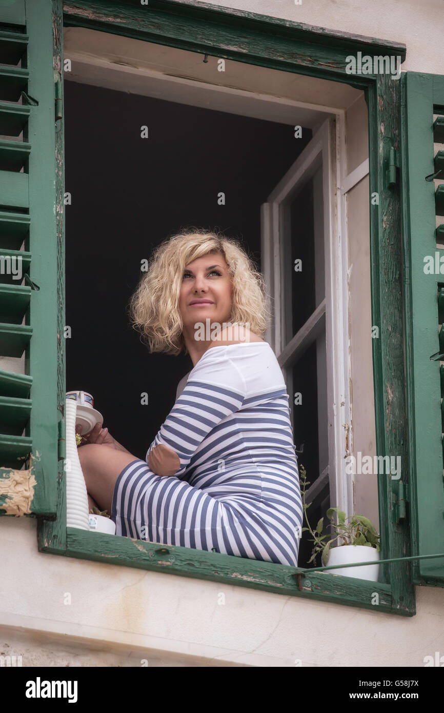 Belle fille en robe rayée avec tasse de café en main est coin sur la fenêtre Banque D'Images
