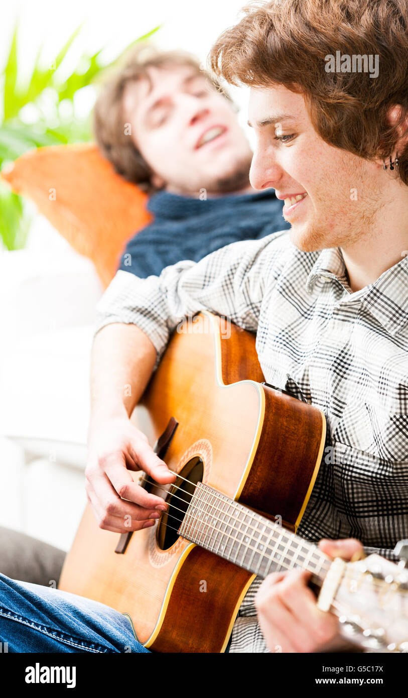 Smiling happy casual young man playing guitar comme son ami se détend sur un canapé derrière lui, appréciant la musique Banque D'Images