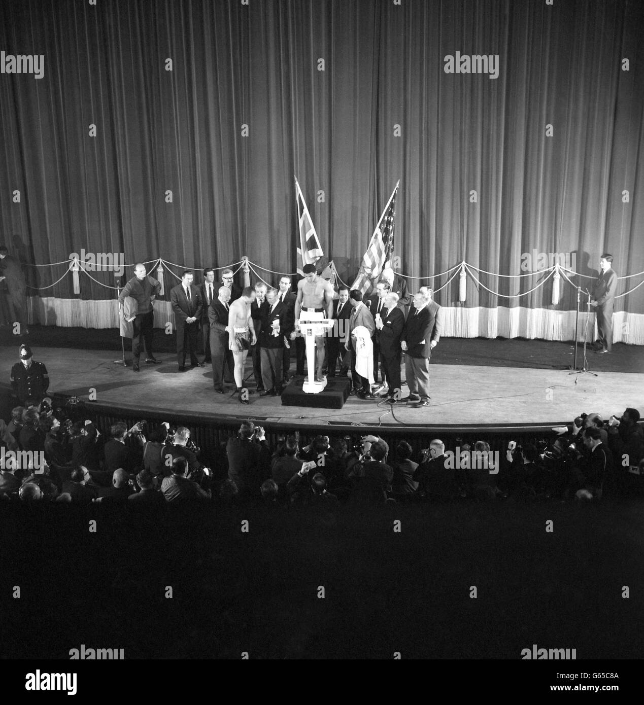 Le poids lourd américain Cassius Clay sur la balance devant les drapeaux américain et britannique, et le challenger britannique Henry Cooper discutant avec Teddy Waltham, secrétaire de la British Boxing Board of Control au Weight-in de l'Odéon à Leicester Square, Londres, pour le combat du Championnat du monde de ce soir. (À gauche) Jim Cooper, qui porte le manteau de son frère, l'entraîneur de Henry Danny Holland et le directeur Jim Wicks, qui ajuste le short du challenger. (À droite) le gérant de Clay, Angelo Dundee (manteau de maintien), le promoteur Harry Levene (programme de maintien) et Micky Duff (à l'épaule droite de Levene). Banque D'Images