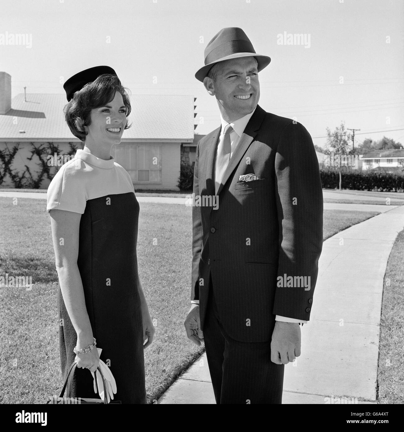 1960 SMILING COUPLE WEARING ROBE VÊTEMENTS DEBOUT SUR UN TROTTOIR DU QUARTIER Banque D'Images