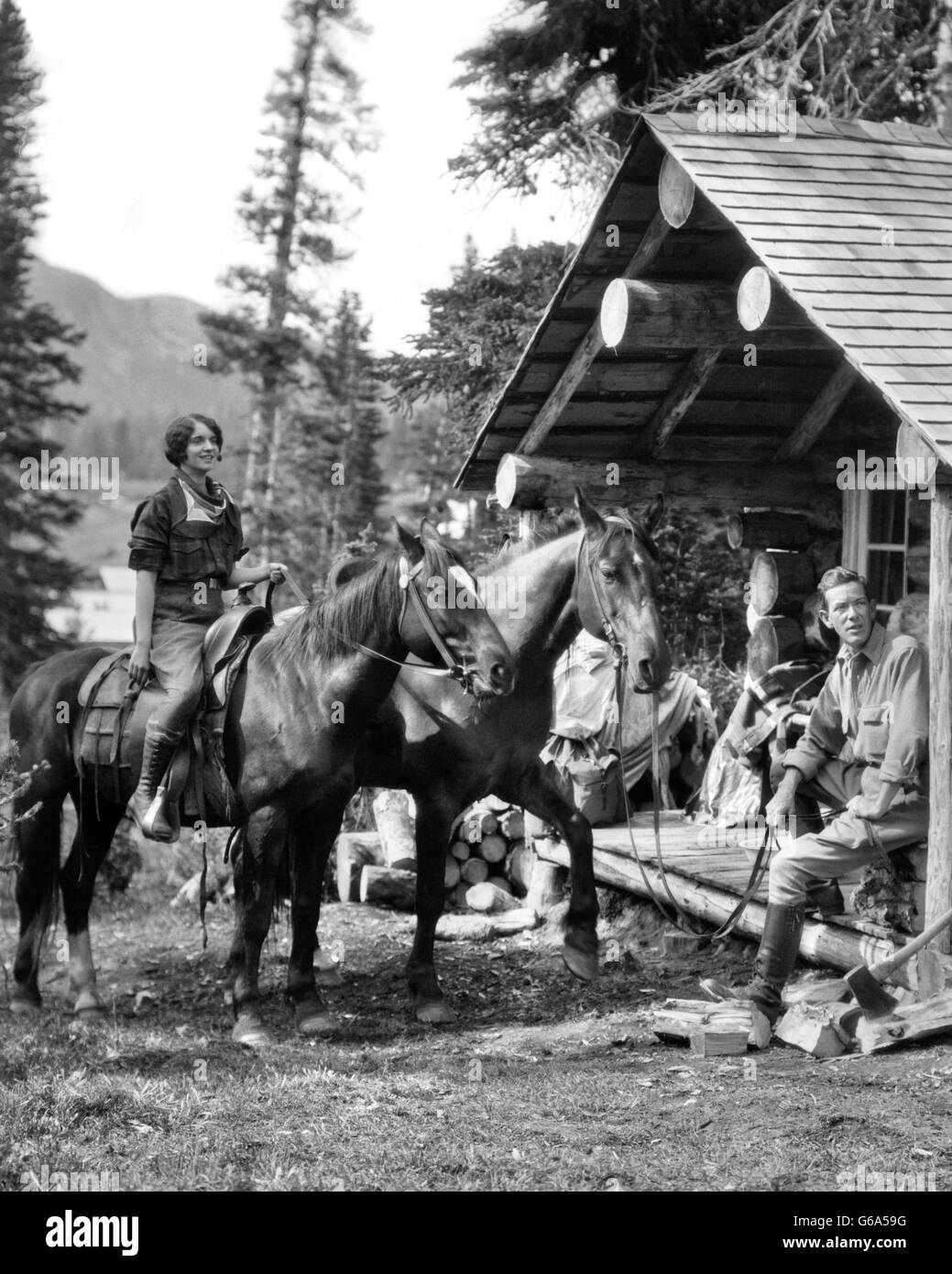 1930 COUPLE IN FRONT OF LOG CABIN FEMME SUR L'HOMME ASSIS SUR LE PORCHE HOLDING RÊNES DU DEUXIÈME CHEVAL CANADA ASSINIBOINE Banque D'Images