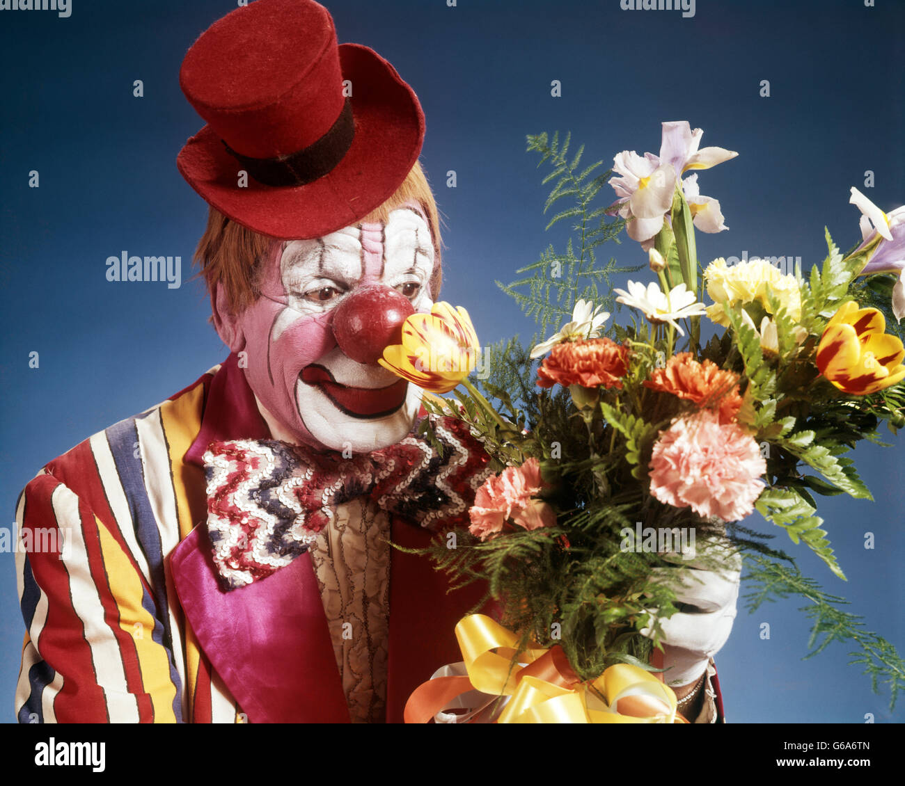 1970 PORTRAIT HAPPY clown de cirque portant chapeau rouge minuscule costume rayé noeud papillon grand bouquet de fleurs odorantes Banque D'Images