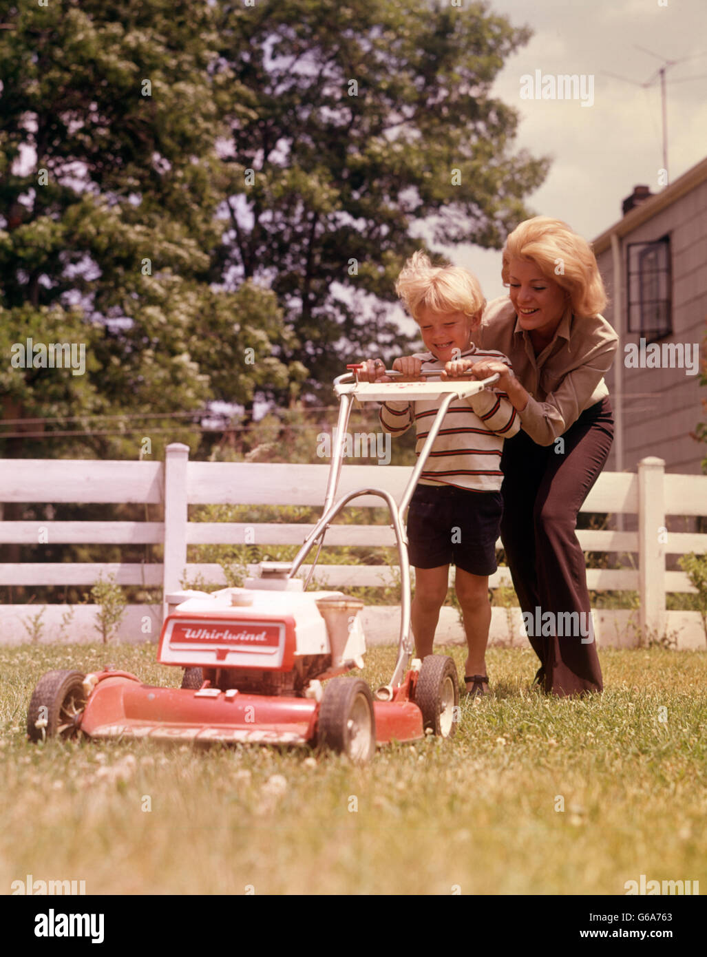 Années 1960 Années 1970 BLONDE MÈRE & son arrière-cour par clôture blanche poussant tondeuse à gazon coupe Banque D'Images