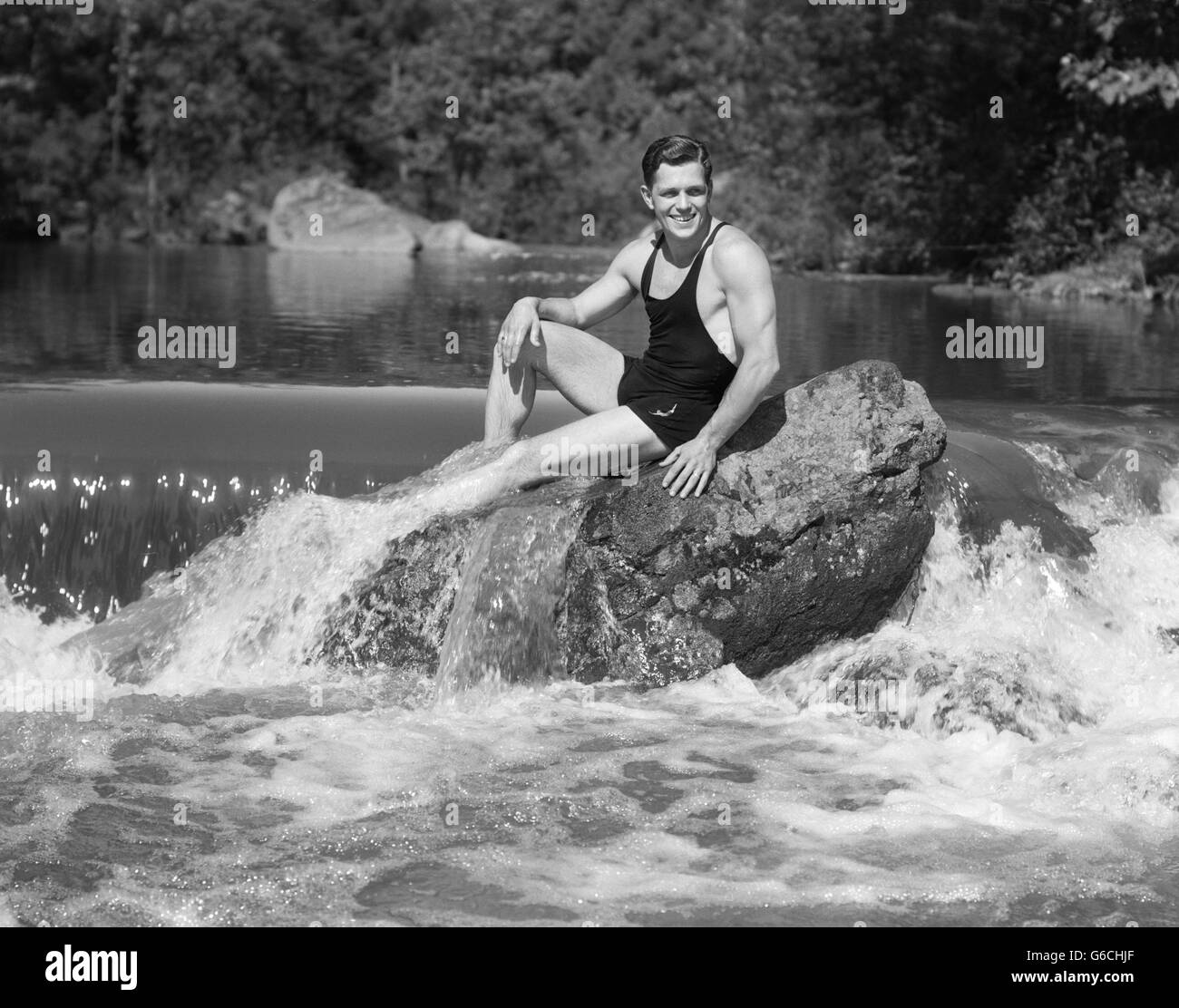 1930 SMILING MAN LOOKING AT CAMERA WEARING BATHING SUIT TANK TOP Short de bain POSANT ASSIS SUR LE GROS ROCHER DANS L'ÉTANG DES BOIS Banque D'Images