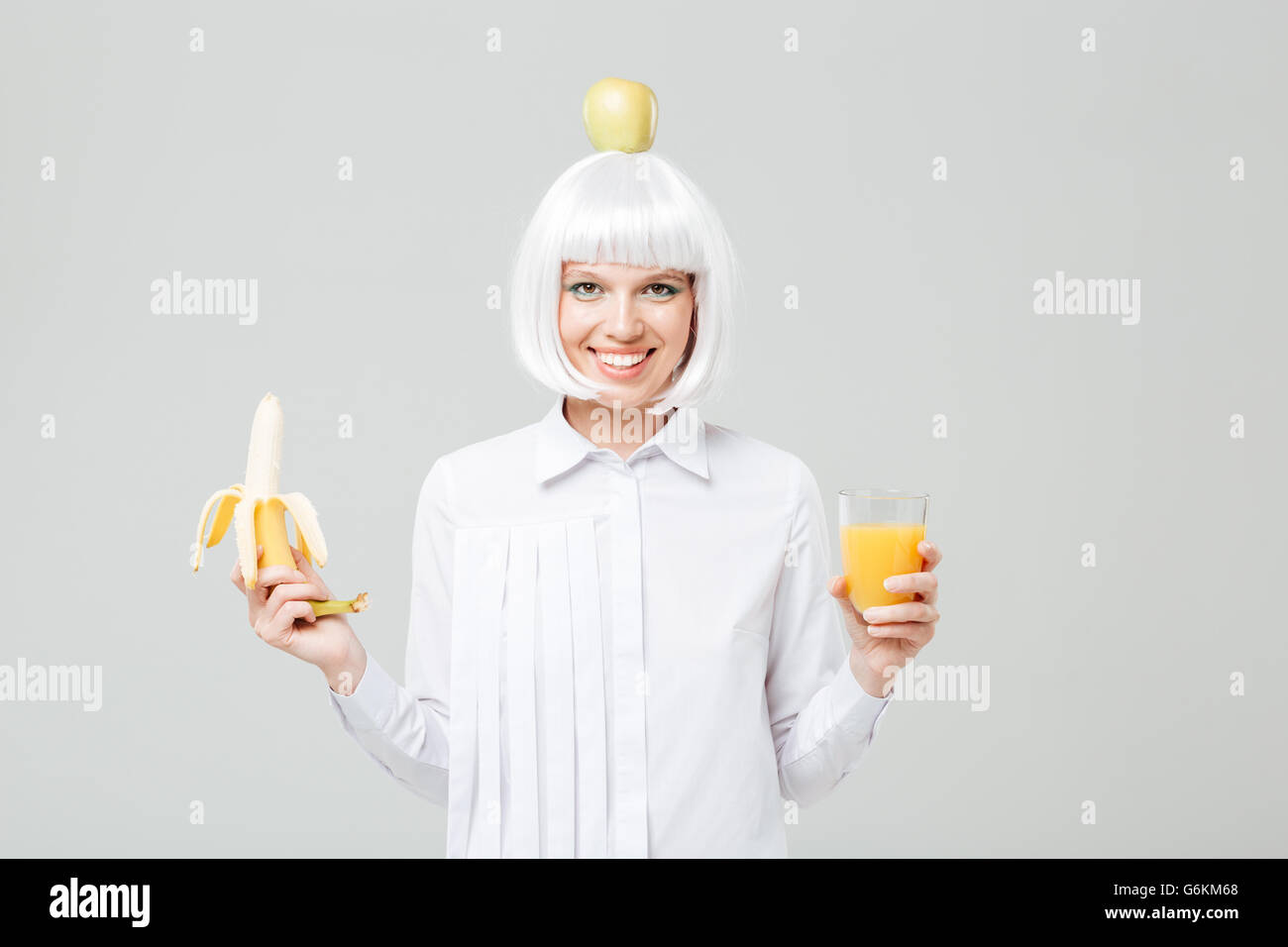 Cheerful young woman with apple sur sa tête holding banana et verre de jus sur fond blanc Banque D'Images