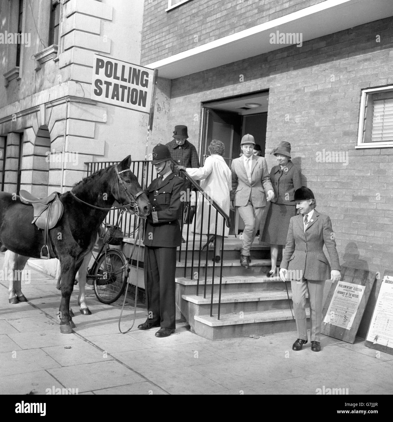 Un policier tient les rênes de deux chevaux, alors que leurs propriétaires terminent leur vote au bureau de vote de la maison Baden-Powell, Queen's Gate, Londres.Les animaux y ont été criés par Dickie Dixon (à droite), directrice de l'école d'équitation de Knightsbridge, et un de ses membres du personnel, René Blick (Steps). Banque D'Images