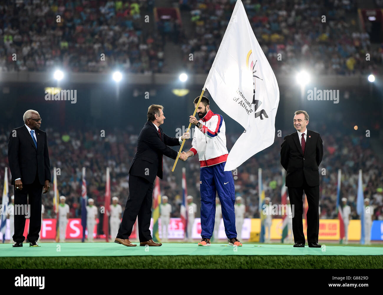 Le capitaine de la Grande-Bretagne Martyn Rooney reçoit le drapeau de la présidente britannique de l'athlétisme Lynn Davies en tant que présidente sortante de l'IAAF Lamine Diack (à gauche) et Simon Cooper, responsable des sports, Bureau du maire de la Greater London Authority, à regarder pendant le neuvième jour des Championnats du monde de l'IAAF au stade national de Beijing, en Chine. Banque D'Images