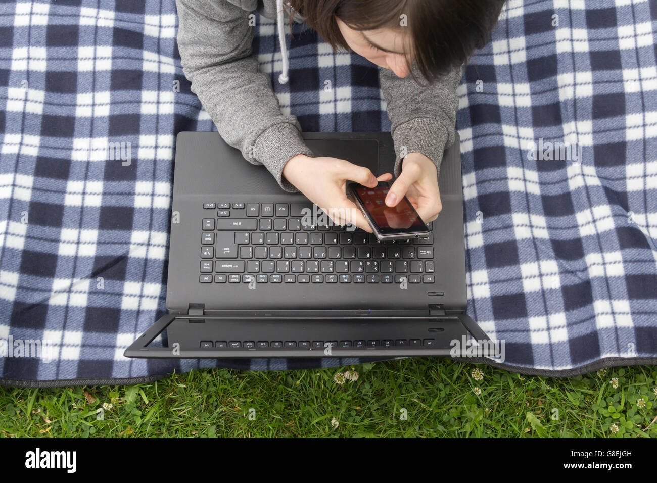 Jeune femme, adulte ou fin de l'adolescence étudiant , allongé sur un tapis de pique-nique sur l'herbe, regardant son smartphone et ordinateur portable. Banque D'Images