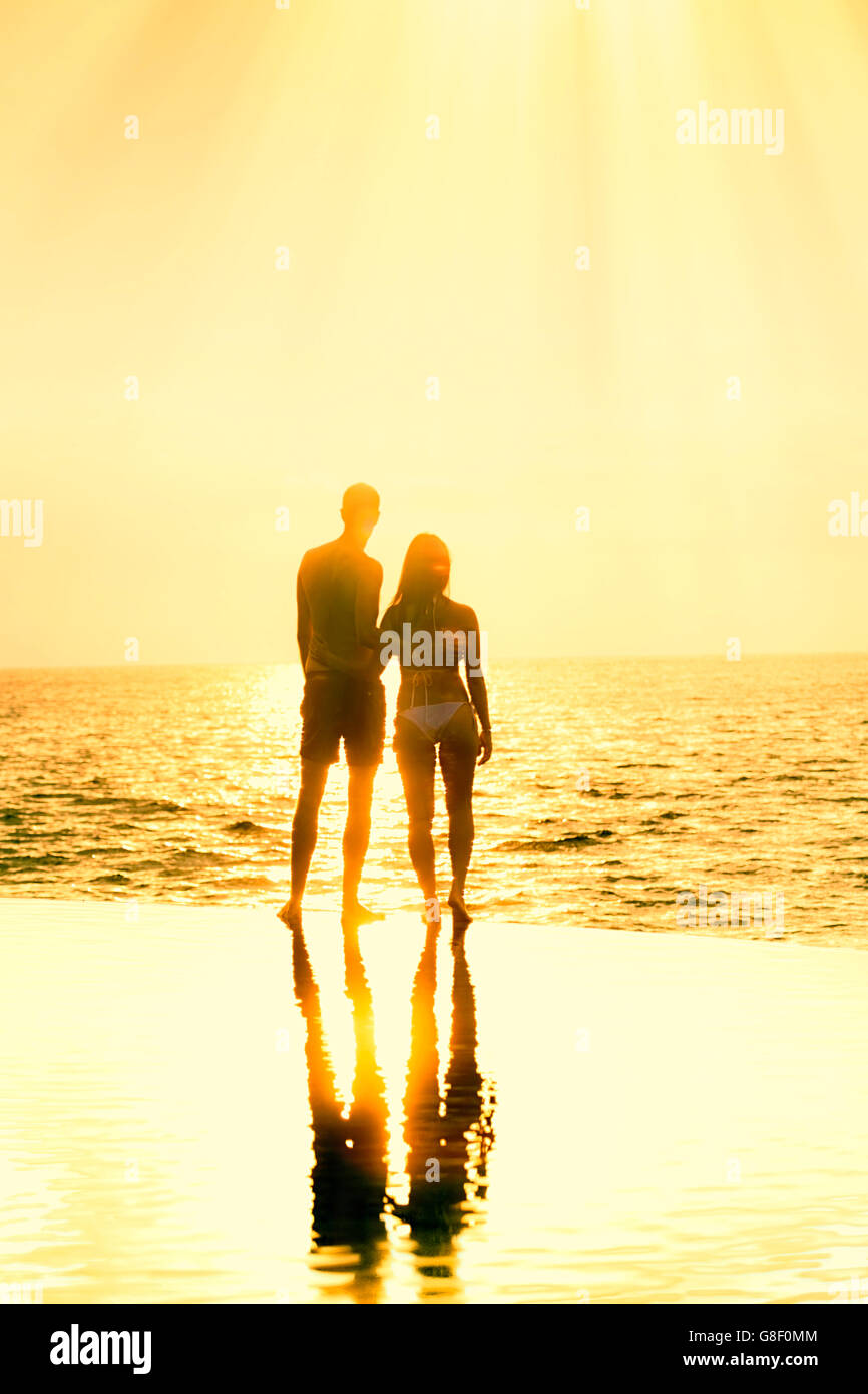Honeymoon couple au bord d'une piscine à débordement à l'encontre d'un coucher du soleil doré/aube Banque D'Images