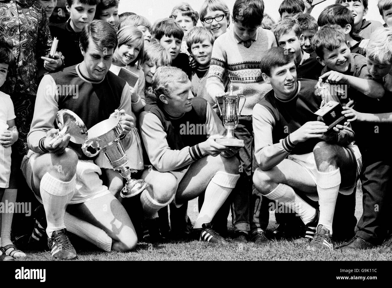 (G-D) le trio de Geoff Hurst, Bobby Moore et Martin Peters, qui a remporté la coupe du monde de West Ham United, examine les trophées avec l'aide de jeunes fans Banque D'Images