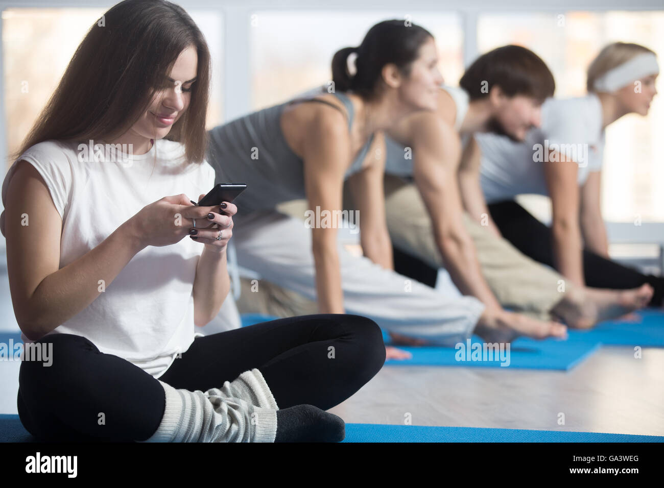 Heureux belle femme jouant avec téléphone au lieu de l'entraînement sportif, assis en tailleur, le repos au cours de la pratique en salle de sport Banque D'Images