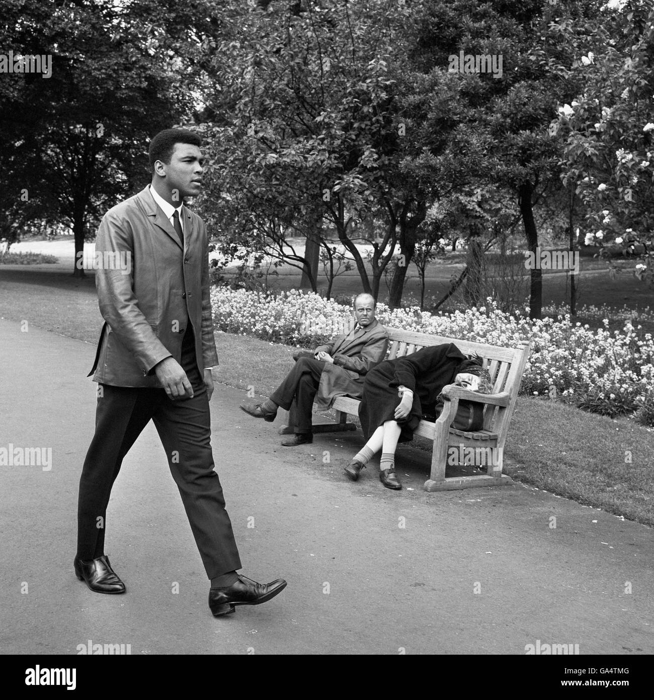 Un Muhammad Ali (Cassius Clay), fraîchement confronté, traverse un parc de Londres le matin après avoir battu le challenger Henry Cooper dans le World Title bout au stade Highbury d'Arsenal. Le combat s'est terminé par un arrêt au 6ème tour pour Muhammad Ali. Deux Londoniens endormis (sur banc) semblent être anonymes à la présence du Champion du monde de poids lourd. Banque D'Images