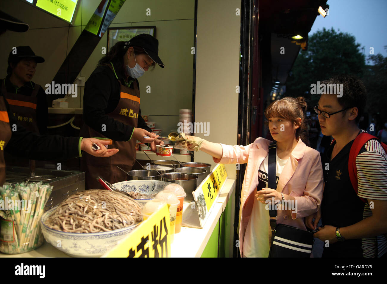 Un homme et une femme s'acheter une tasse de nouilles de fruits de mer à un stand de nourriture. La personne de ventes couvre une face contre la pollution. Beijing, Chine. Banque D'Images