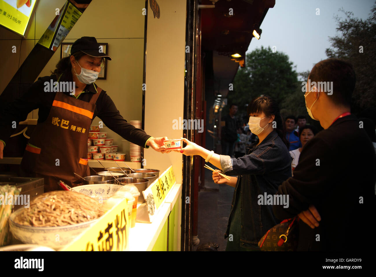 Un homme et une femme s'acheter une tasse de nouilles de fruits de mer à un stand de nourriture. La personne de ventes et ils couvrent leurs visages contre la pollution. Banque D'Images