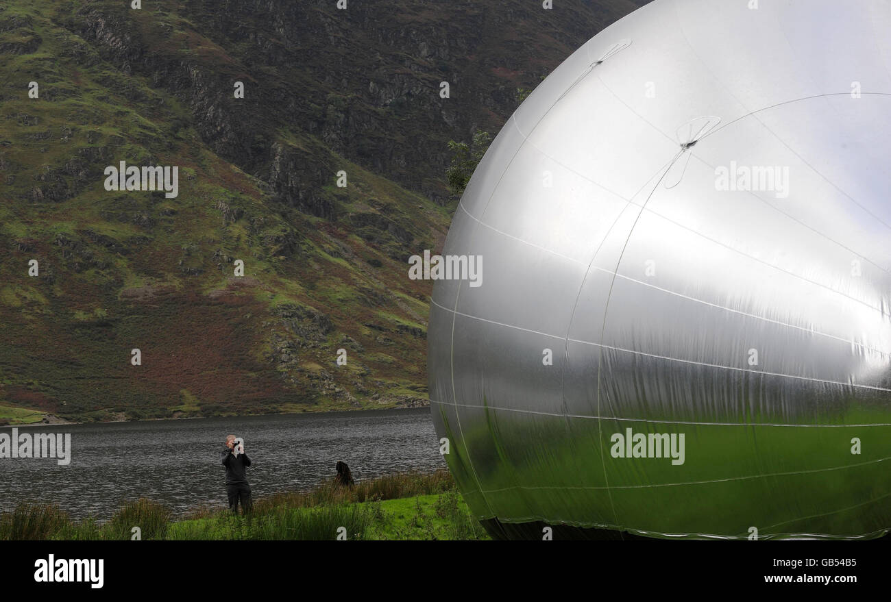 Un marcheur prend une photo de 'drop' une sculpture gonflable réfléchissante géante inspirée d'un raindrop et conçue par l'artiste local Steve Messam est vue sur la rive de Crummock Water dans le Lake District. La sculpture est conçue pour refléter le paysage spectaculaire environnant à l'effet étonnant et est de la taille d'un bâtiment de trois étages. Banque D'Images