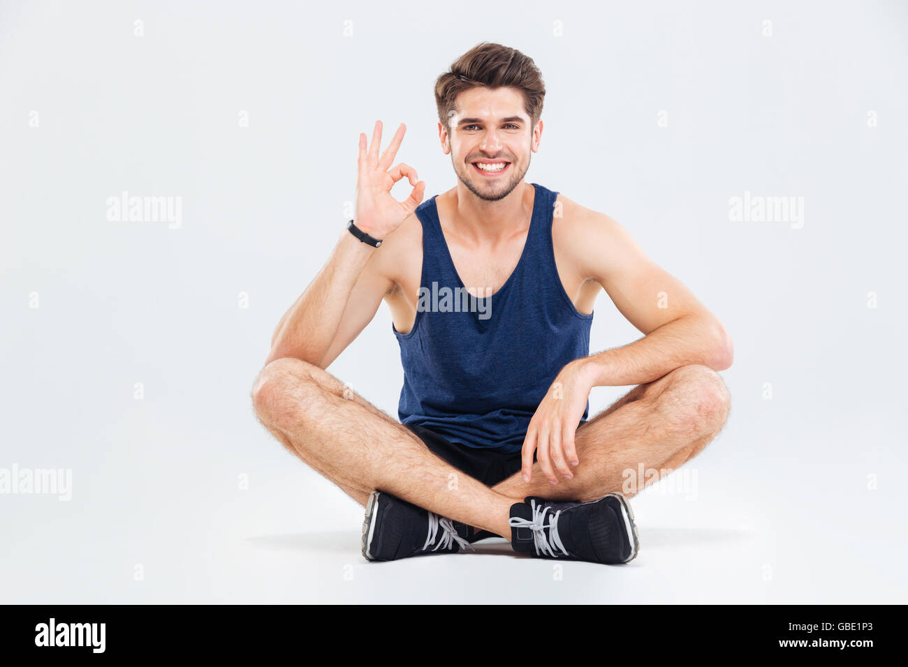 Jeune homme sportif assis avec les jambes croisées et montrant ok sign over white background Banque D'Images