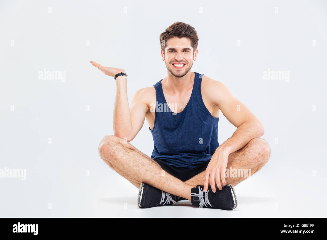 Heureux jeune homme athlétique assis avec les jambes croisées et holding copyspace sur palm sur fond blanc Banque D'Images