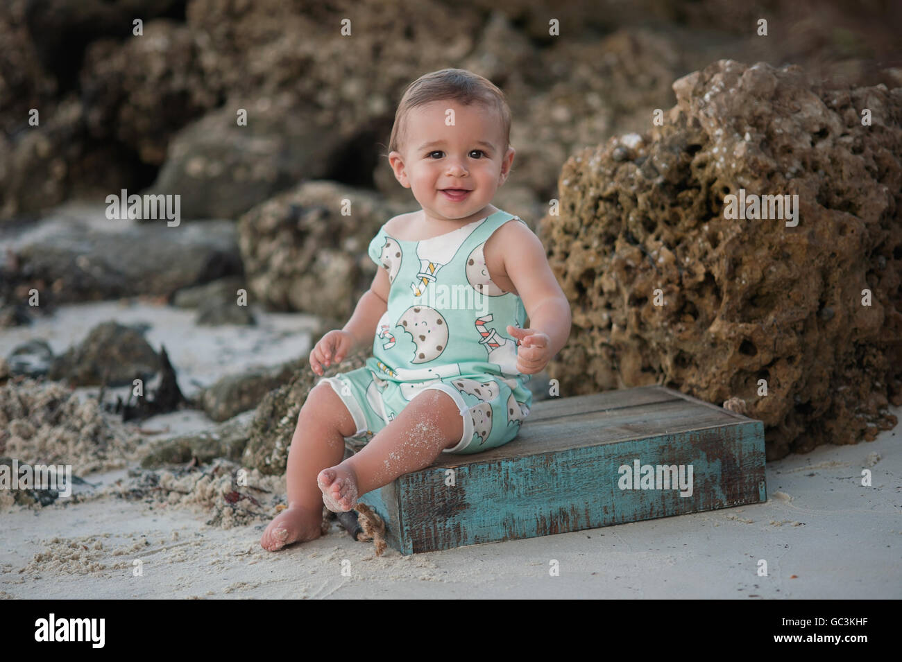 Un petit garçon âgé d'un an, souriant et assis sur une caisse en bois. Tourné en extérieur sur une plage de rochers à l'arrière-plan. Banque D'Images