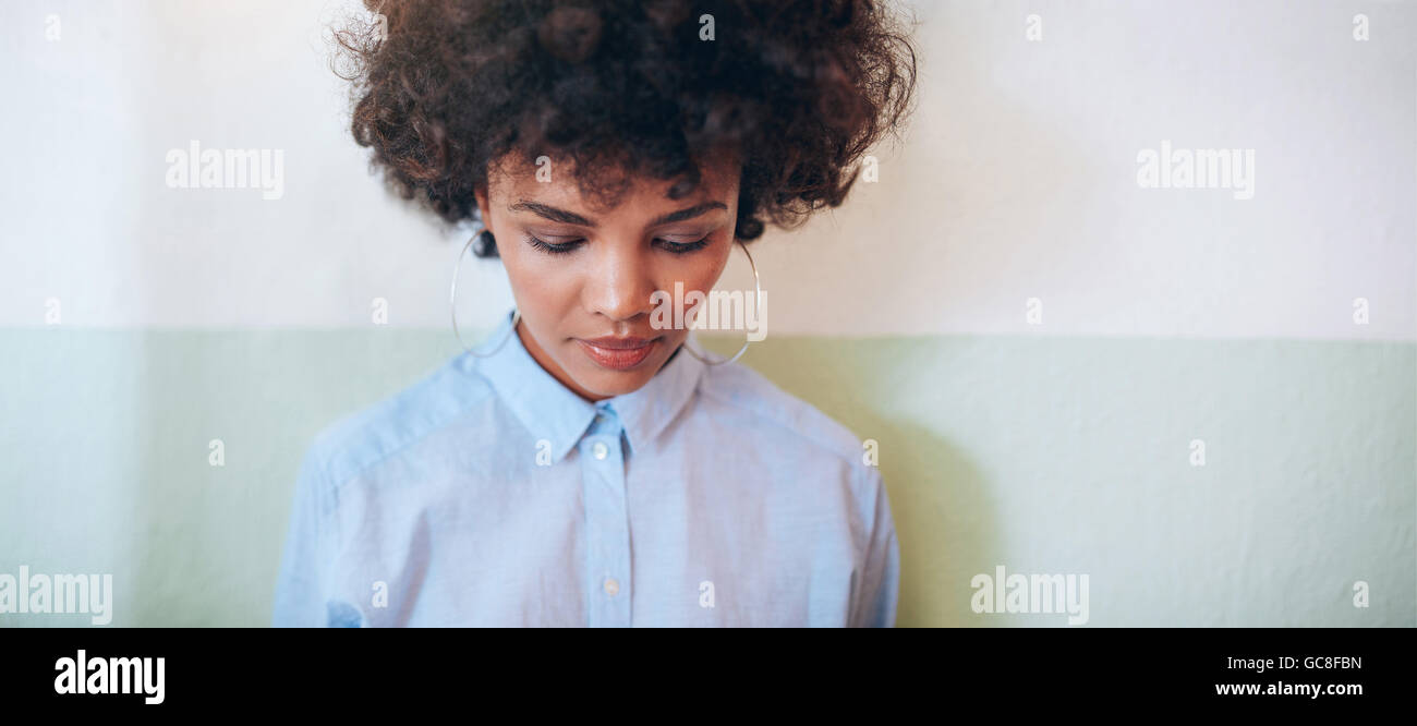 De l'horizontale avec les cheveux bouclés à la recherche vers le bas en position debout contre un mur. African woman looking pensive. Banque D'Images