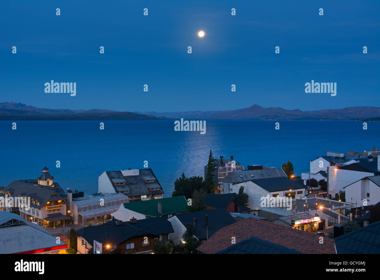 La Lune se levant au-dessus d'un lac au-dessus de la ville de toits montrant quelques lumières de la ville jaune sur le fond bleu, Bariloche, Argentine Banque D'Images