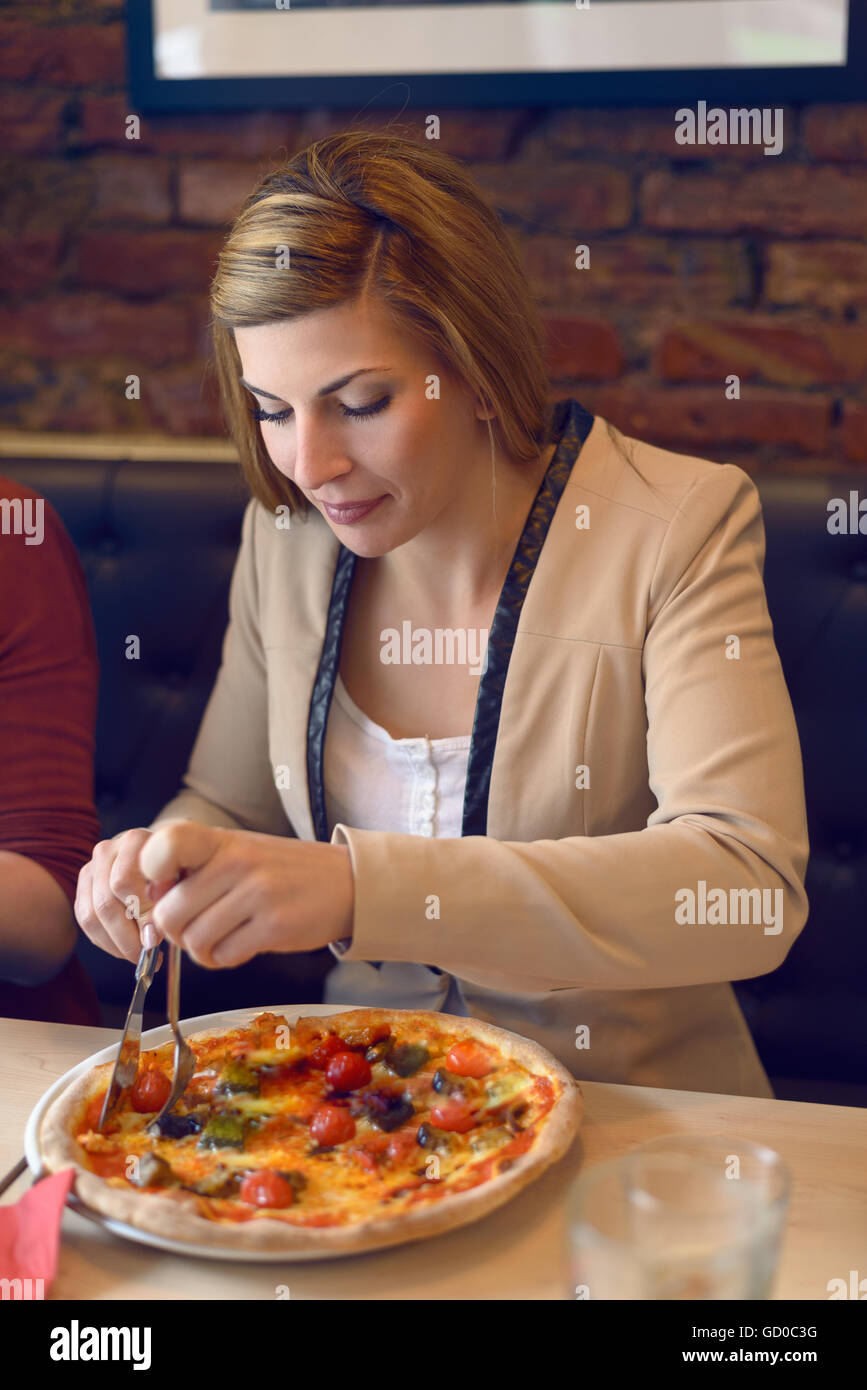 Happy young woman in veste marron et de longs cheveux blonds de manger une délicieuse pizza italienne à table in restaurant Banque D'Images