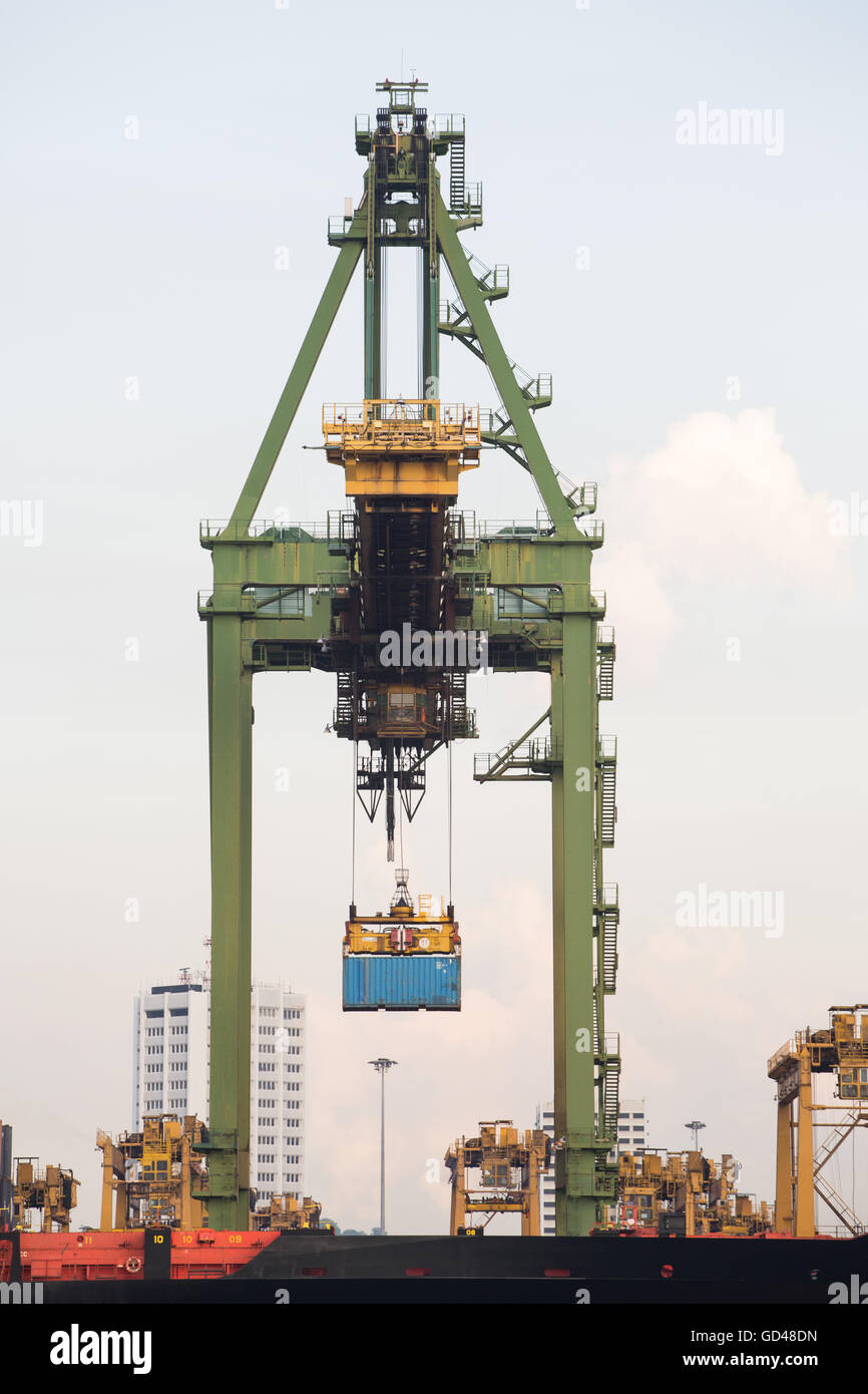 Une perspective de vue frontale verticale d'une grue à conteneurs industrielle en cours de traitement soulevant un conteneur bleu au terminal portuaire. Singapour. Banque D'Images