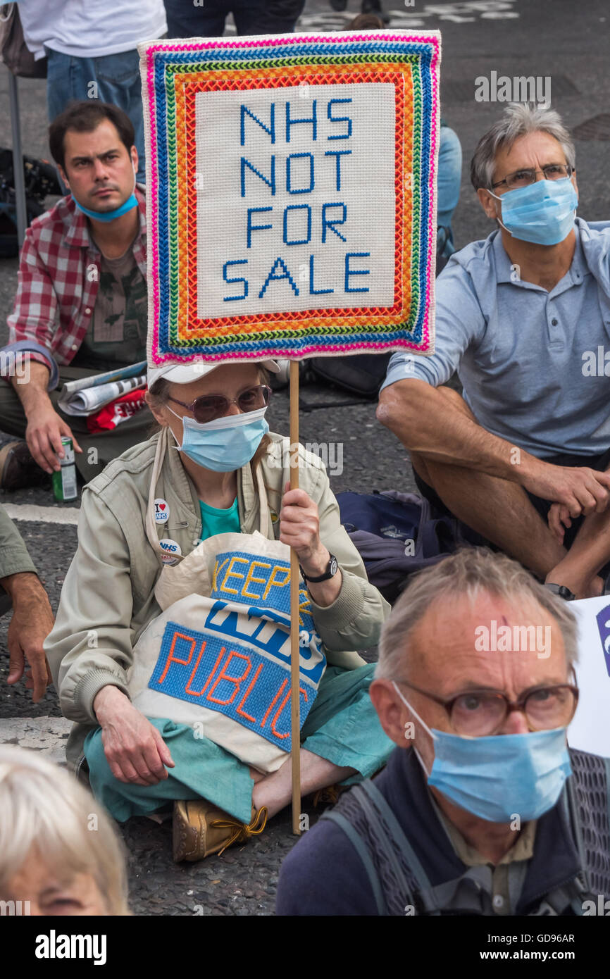 Londres, Royaume-Uni, le 14 juillet 2016. Asseoir des manifestants portant des masques chirurgicaux avant le mois de mars à St Pauls. Une femme montre une crocehted placard 'NHS NI À VENDRE' . L'événement était organisé par la solidarité du NHS, les travailleurs de la santé et de supports dédiés à la sauvegarde d'un système public, livré et responsable NHS . Peter Marshall/Alamy Live News Banque D'Images