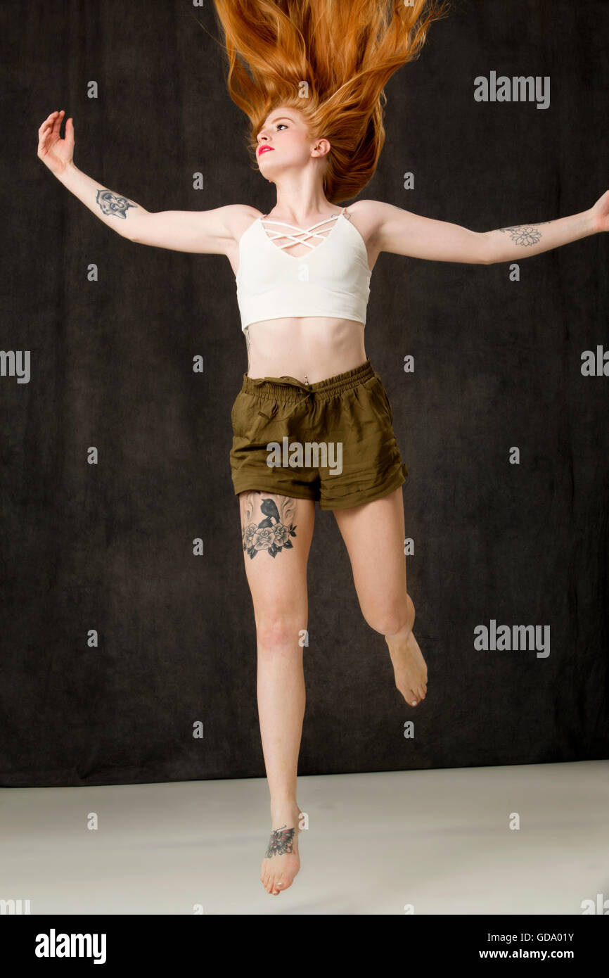 Jeune femme aux longs cheveux rouges et des tatouages portant des shorts vert l'atterrissage d'un saut, cheveux vers le haut. Banque D'Images
