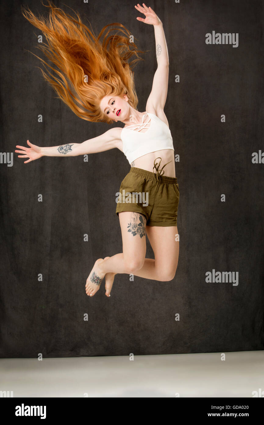 Jeune femme aux longs cheveux rouges et des tatouages portant des shorts vert et sautant dans les airs avec le streaming de cheveux derrière. Banque D'Images