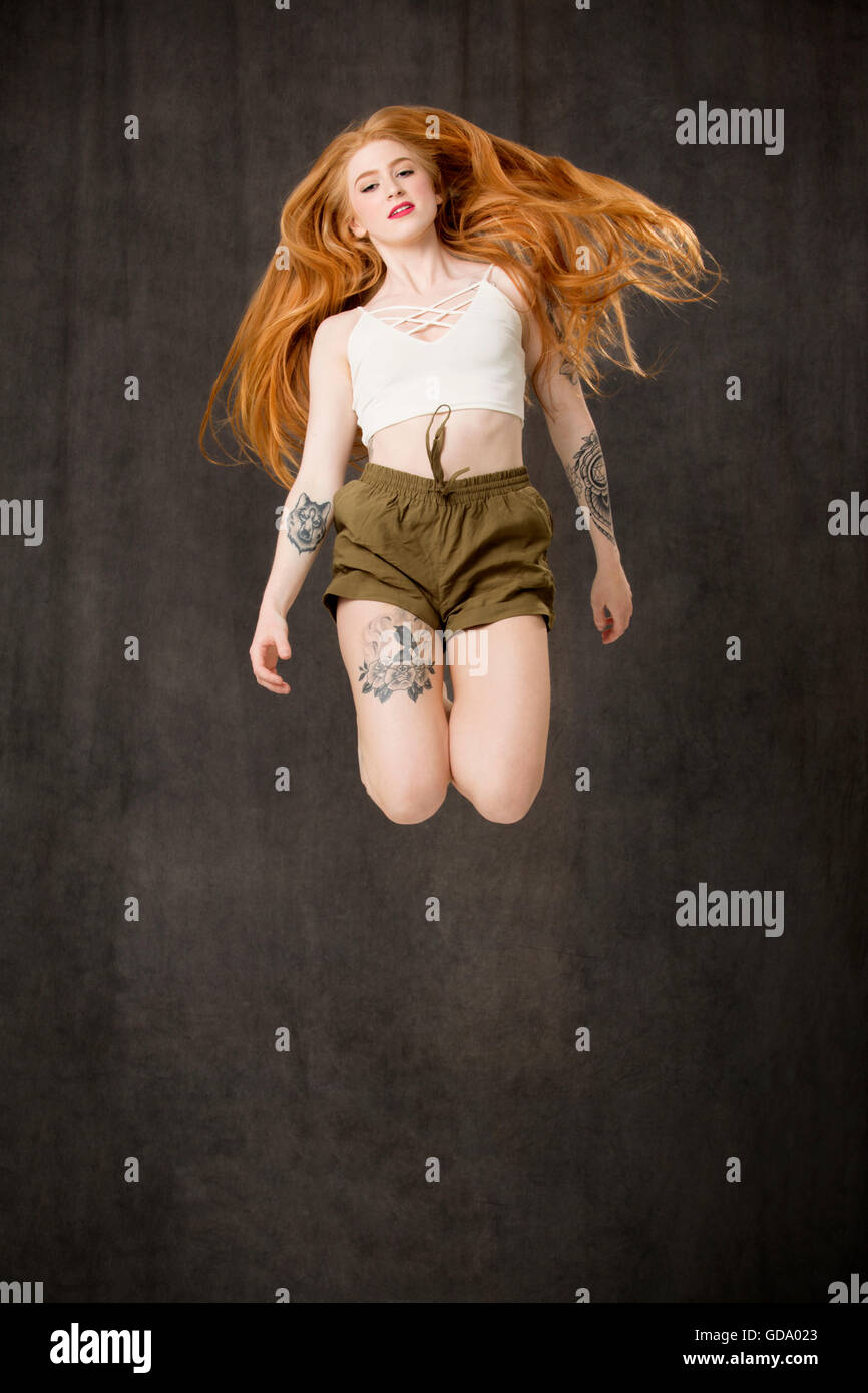 Jeune femme aux longs cheveux rouges et des tatouages portant des shorts vert et sautant à pieds et les cheveux sur les épaules. Banque D'Images