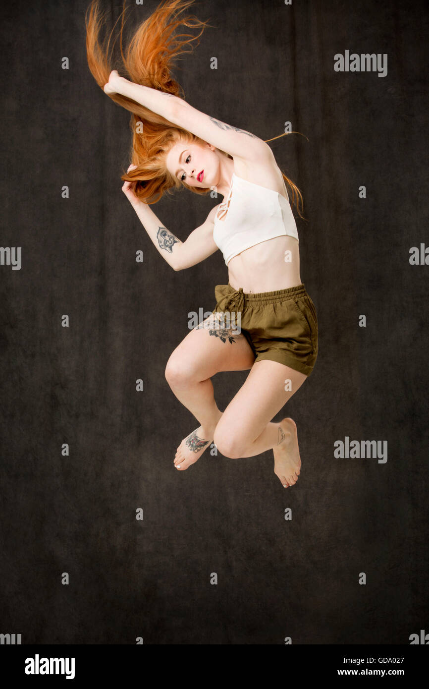 Jeune femme aux longs cheveux rouges et des tatouages portant des shorts vert et sauter avec les genoux pliés et les bras. Banque D'Images