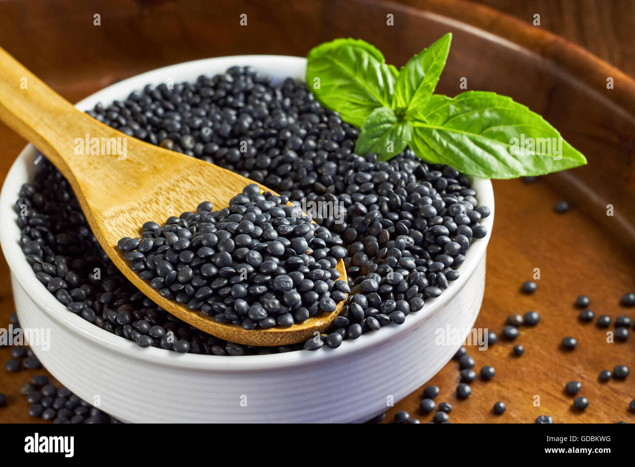 Bol blanc rond plein de lentilles béluga et les feuilles de basilic avec une cuillère de bois sur fond de bois brun. Banque D'Images