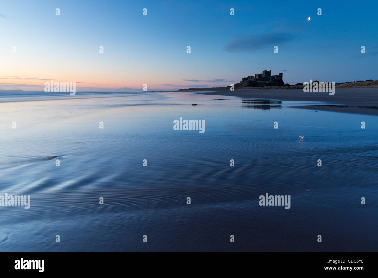 Le château de Bamburgh se reflète en partie dans le sable humide de la plage tandis que les vagues se retatent, la plage de Bamburgh Banque D'Images