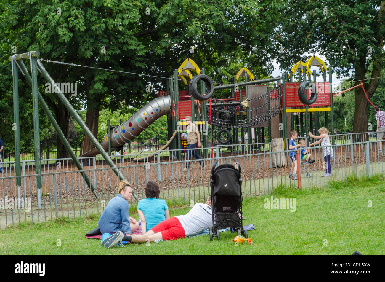 Les parents se relaxent sur l'herbe pendant que leurs enfants jouent dans l'aire de jeux dans la région de Prospect Park, lecture. Banque D'Images