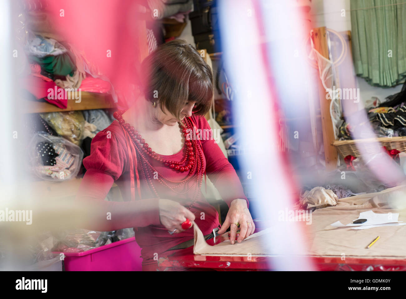 Une couturière en blouse rouge et colliers mesure et coupe de tissu blanc sur la table de coupe avec des rubans au premier plan. Banque D'Images