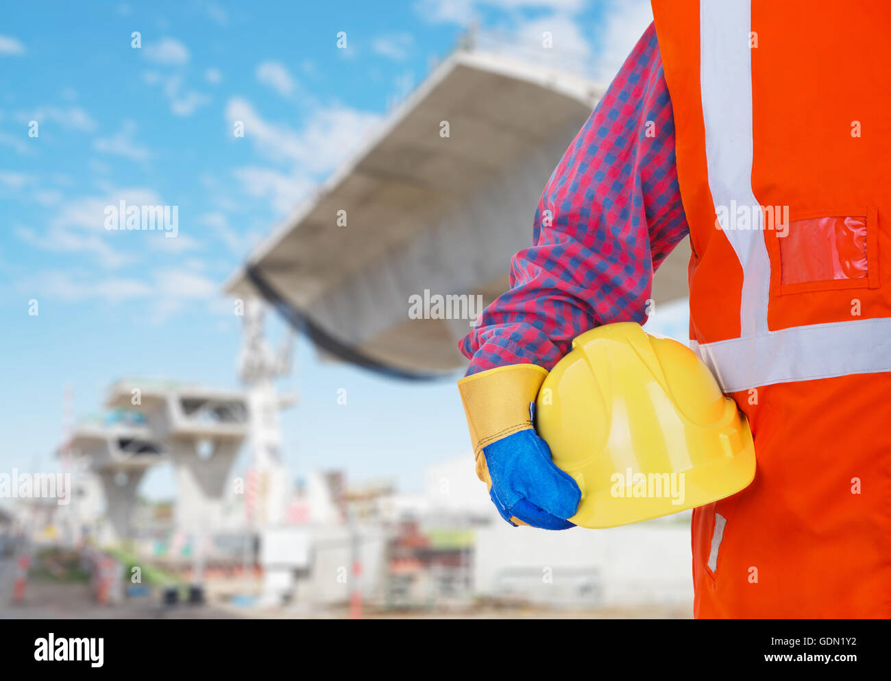 De Protection de la sécurité des équipements de travail. Travailleur en gilet orange holding casque jaune contre la construction en arrière-plan du site Banque D'Images