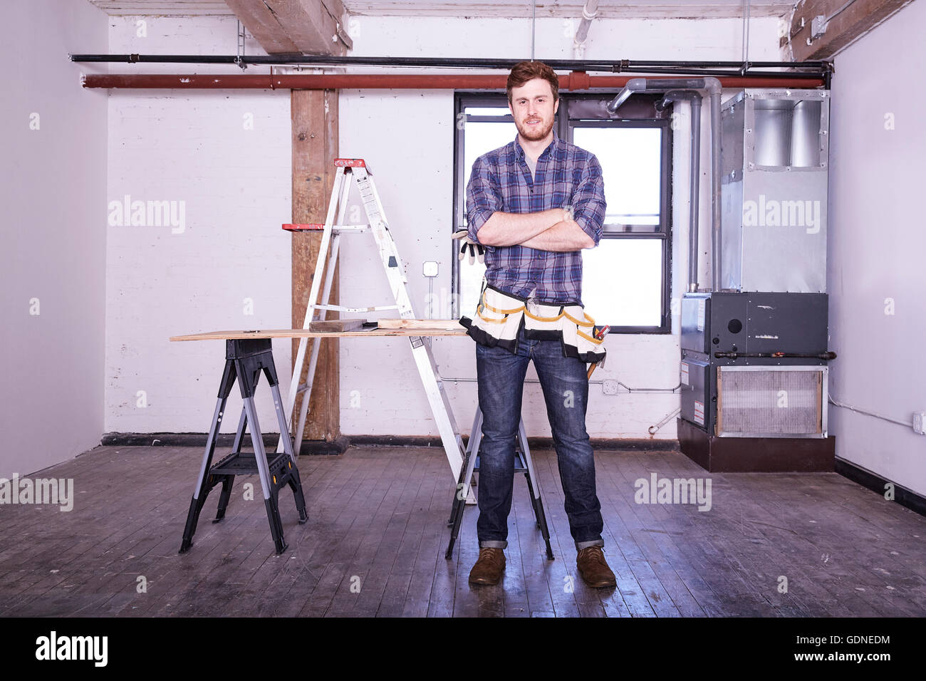 Jeune homme en atelier wearing tool belt, bras croisés looking at camera Banque D'Images