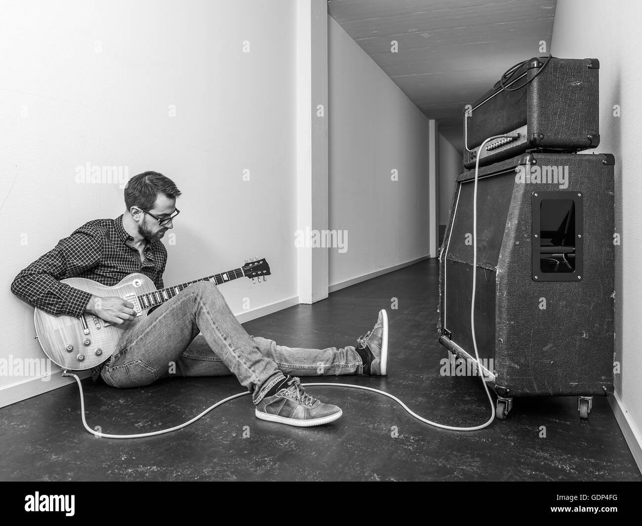 Photo d'un homme assis jouant de sa guitare électrique en face d'un grand amplificateur dans un couloir. Version noir et blanc. Banque D'Images