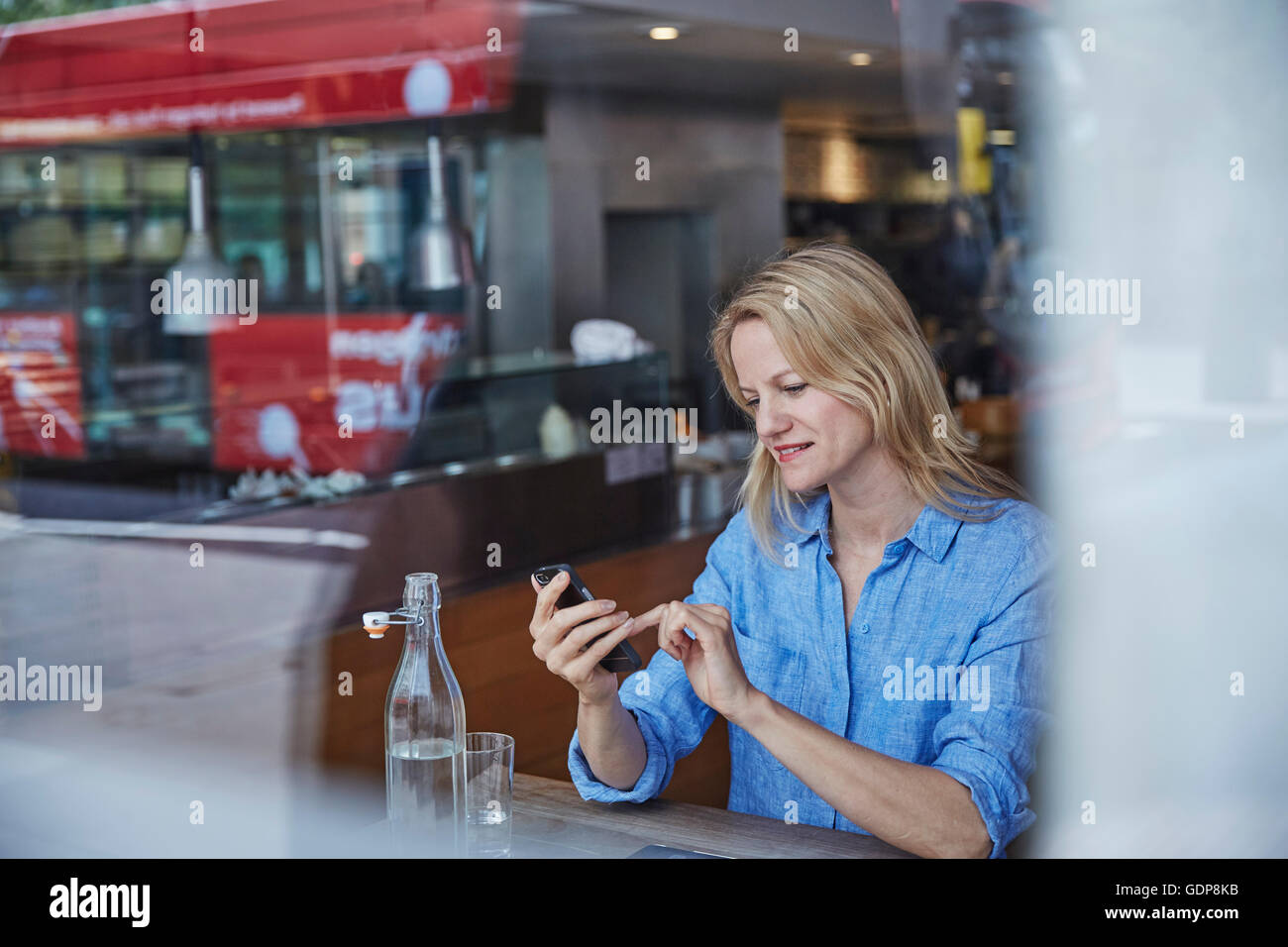Mature Woman sitting in cafe, using smartphone, bus reflète dans window Banque D'Images