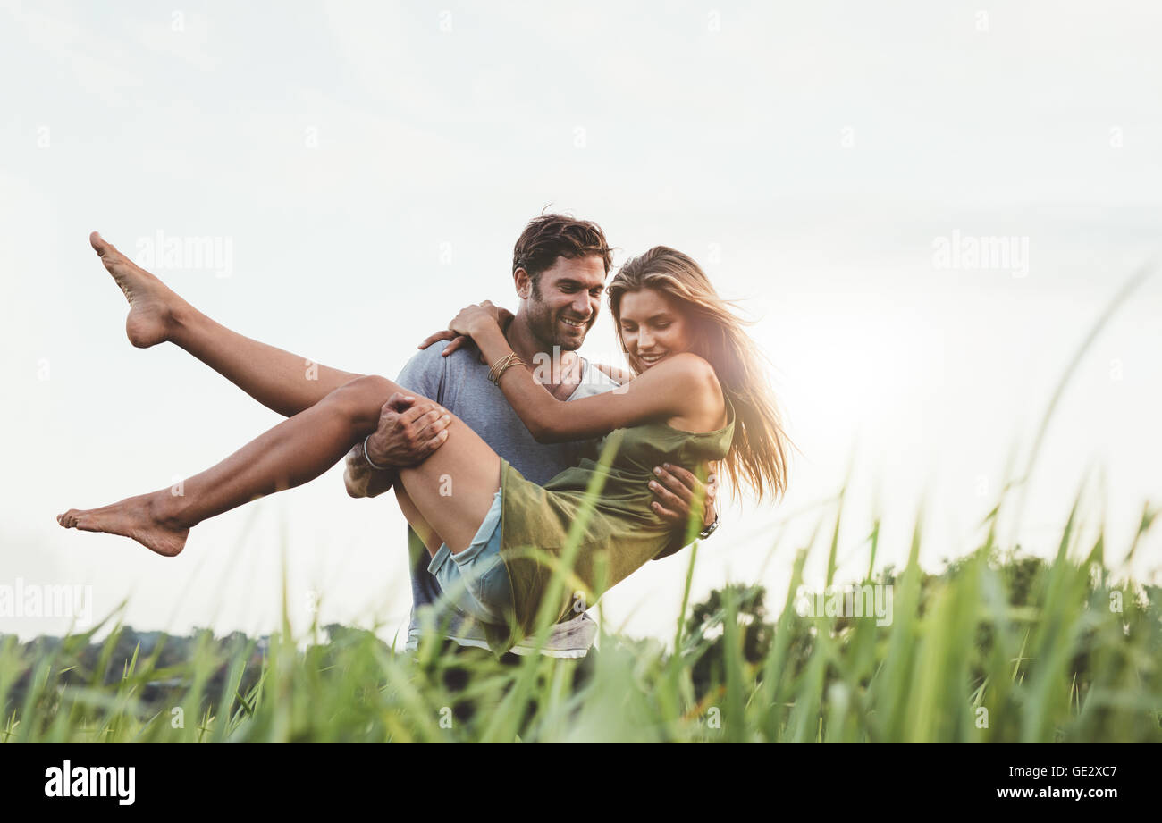 Coup de jeune homme portant sa belle petite amie dans les bras et marche à travers champ d'herbe. Couple dans la nature sur un somm Banque D'Images