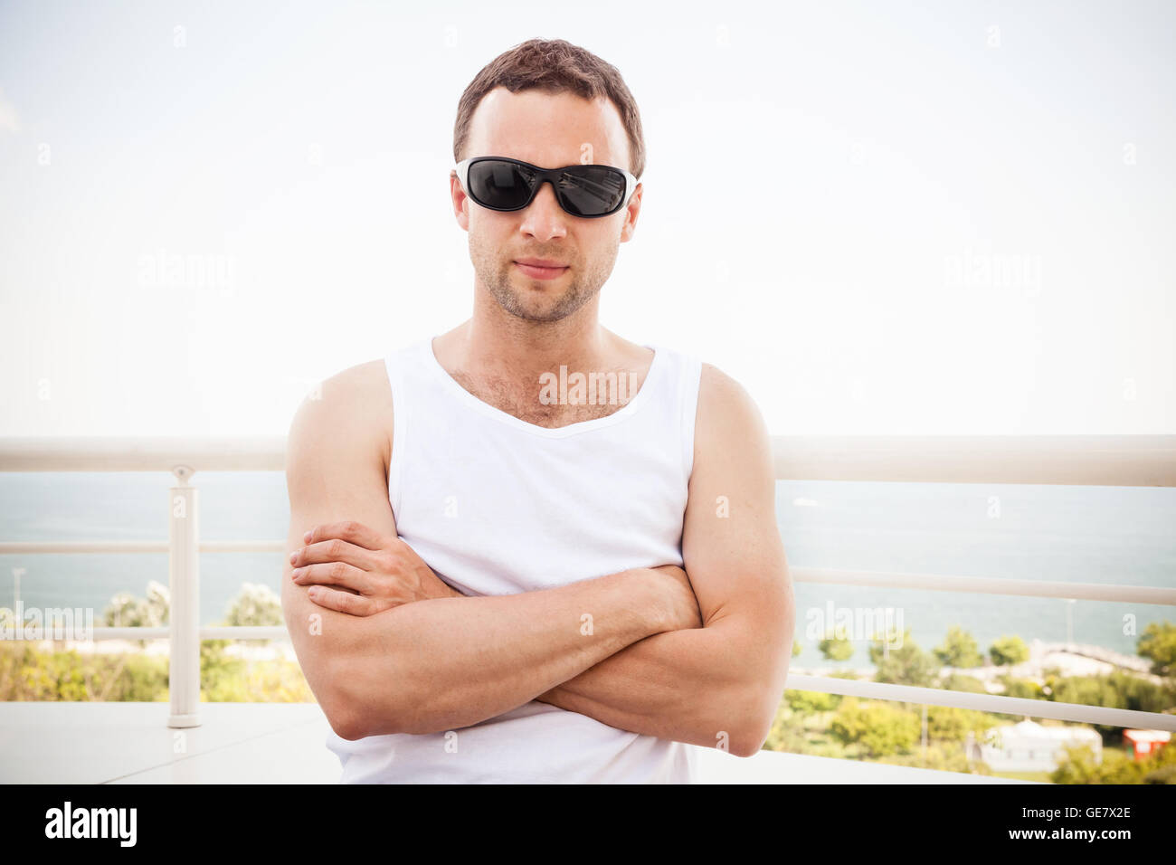 Young Caucasian man en chemise blanche et lunettes noires se tient avec les mains de passage. Portrait d'été en plein air avec vue mer sur un Banque D'Images