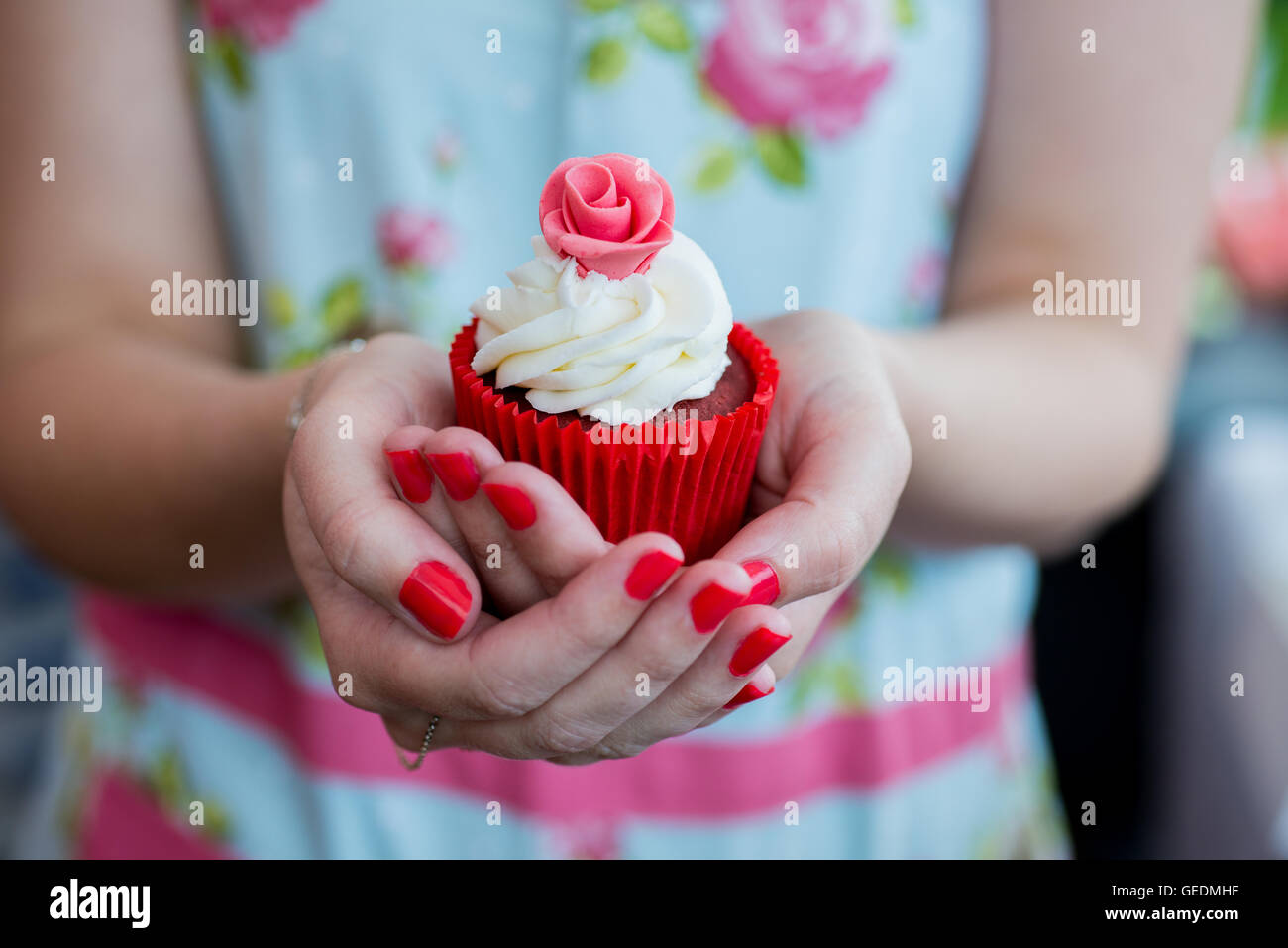 Close up shot of a womans mains tenant un joli cupcake. Elle porte une fleur lumineuse robe la réécriture. Le cupcake est en rouge une p Banque D'Images