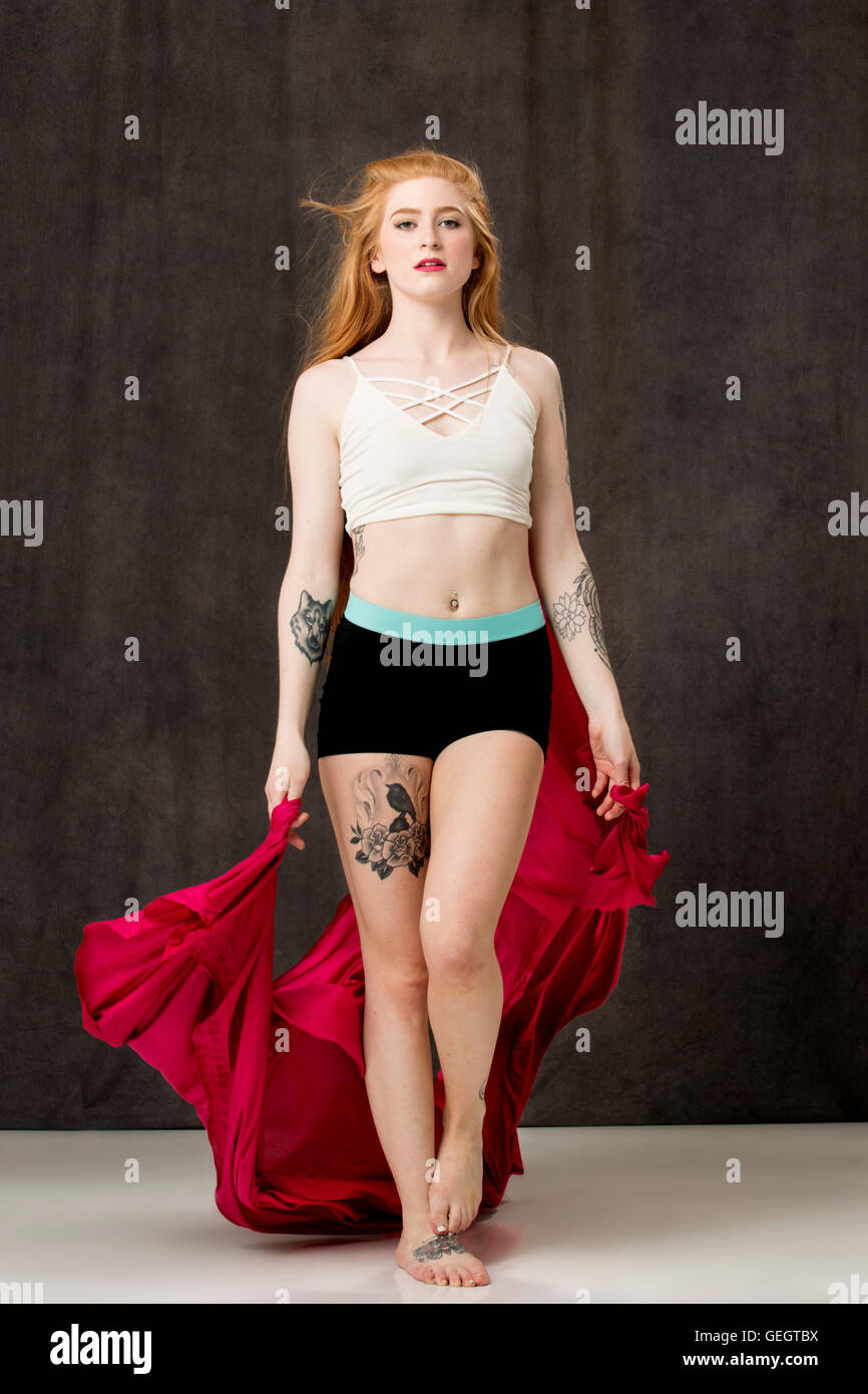 Jeune danseuse avec de longs cheveux rouges et tatouages, tissu rouge holding que les sentiers derrière elle alors qu'elle marche en avant. Banque D'Images