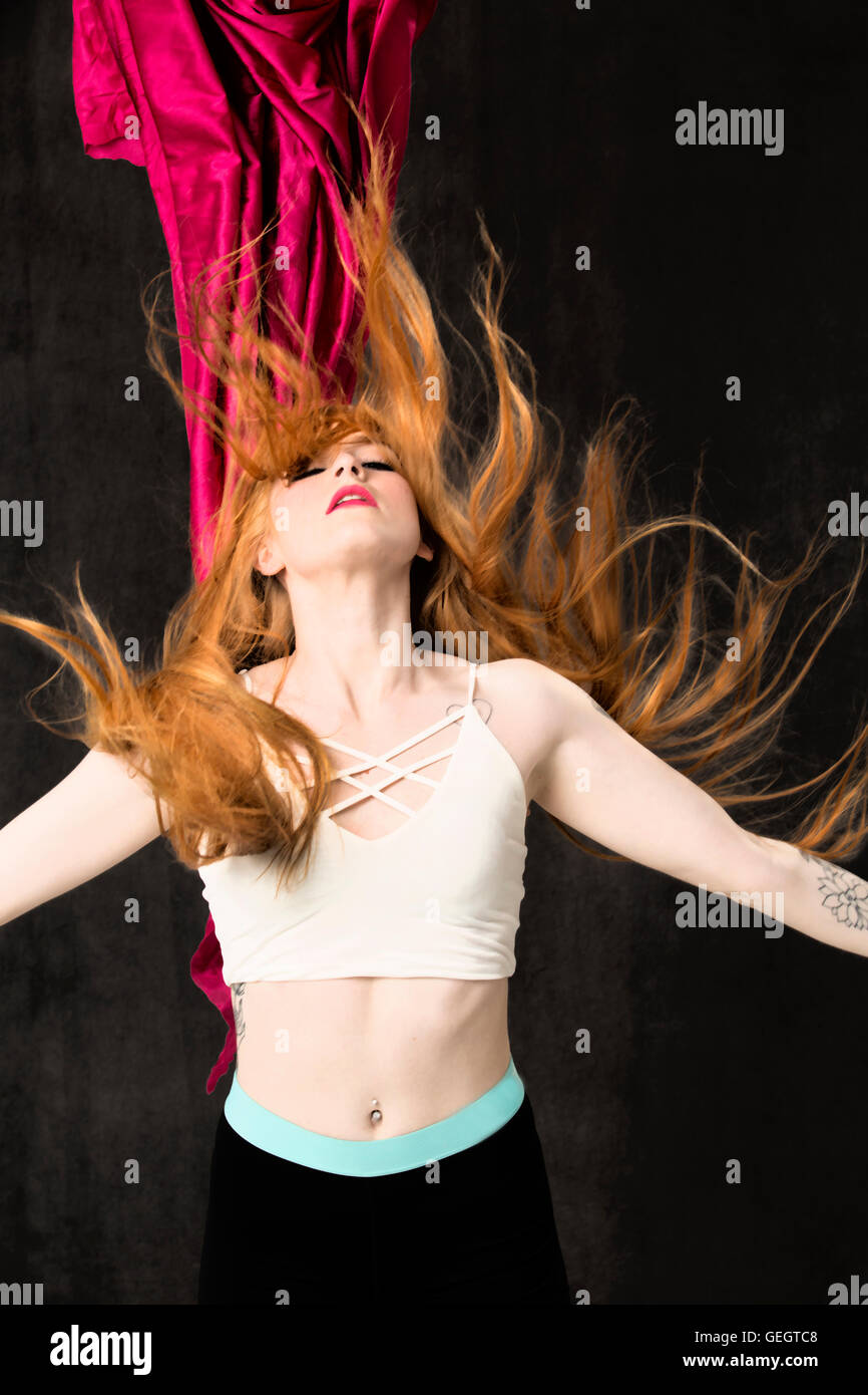 Jeune danseuse aux longs cheveux rouges et des tatouages dont les traces de cheveux vers le haut comme les flammes, avec tissu rouge suspendue derrière elle. Banque D'Images
