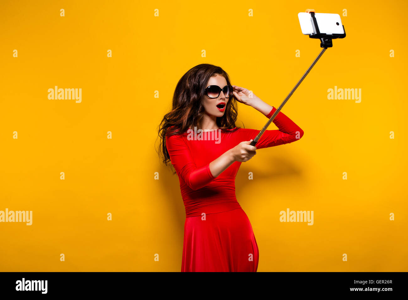 Portrait de la belle brune en robe rouge lèvres impressionnant et portant des lunettes de soleil tout en faisant avec selfies stick. Isolé Banque D'Images